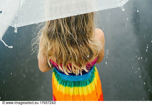 Blond girl is under and umbrella in rain storm in rainbow dress