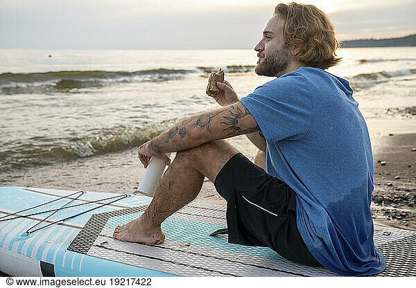 Blond man eating sandwich sitting on paddleboard at beach