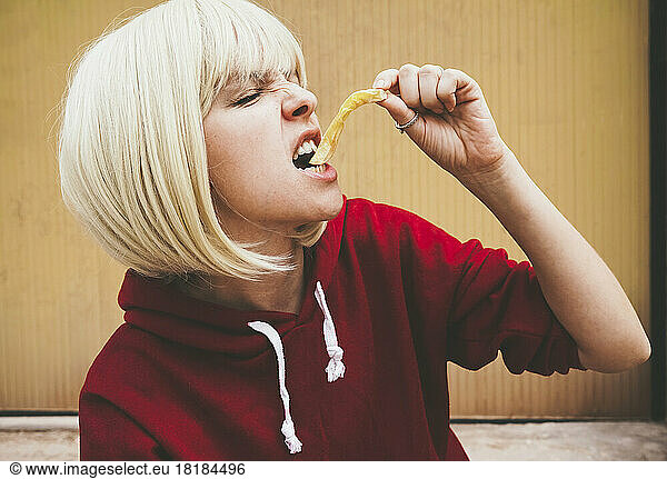 Blond woman eating French fries in front of brown wall