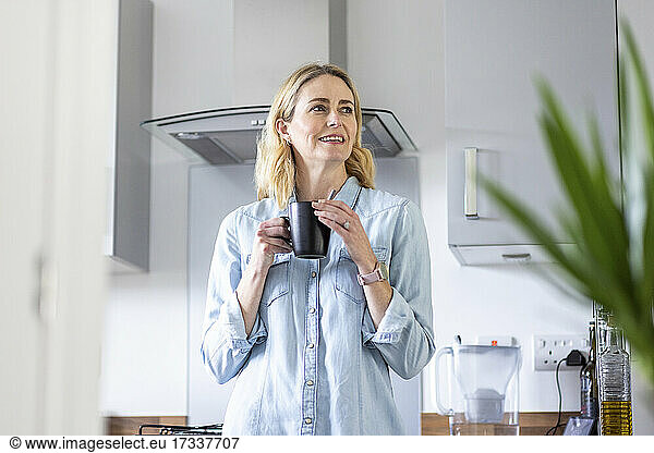 Blond woman holding mug while looking away in kitchen at home