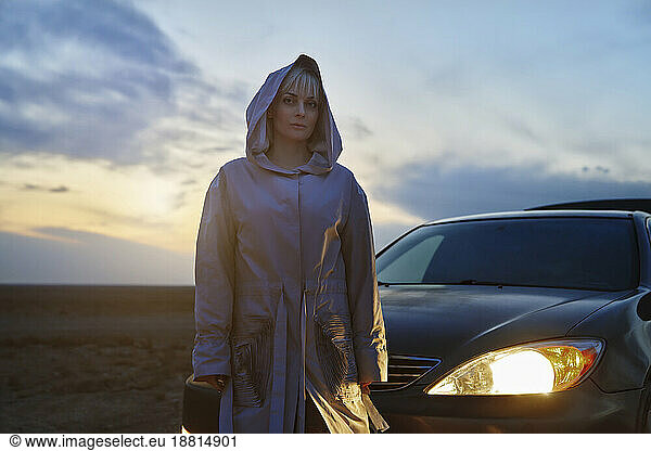 Blond woman in raincoat standing next to illuminated headlight of car
