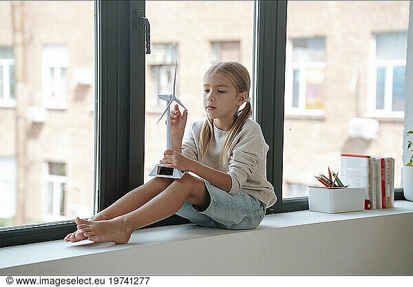 Blond woman playing with wind turbine model on window sill at home