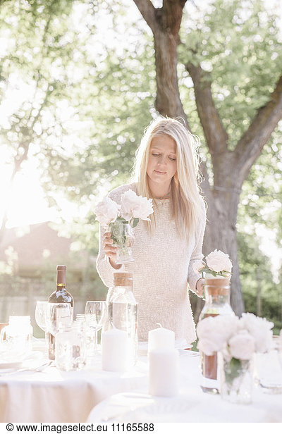 Blond woman setting a table in a garden  candles and vases with pink roses.