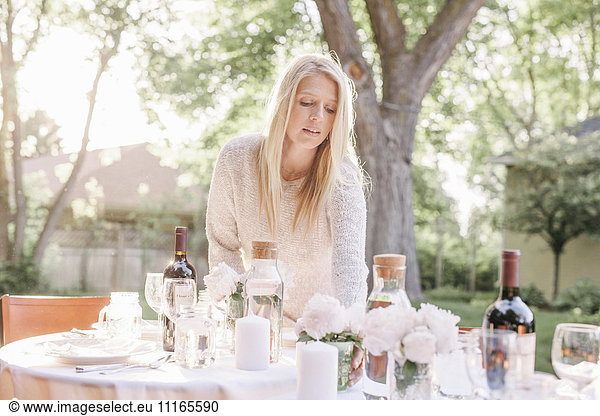 Blond woman setting a table in a garden  vases with pink roses.