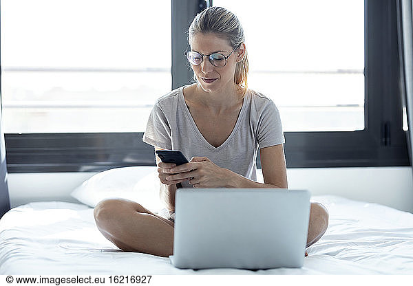 Blond woman using laptop and smartphone sitting on bed