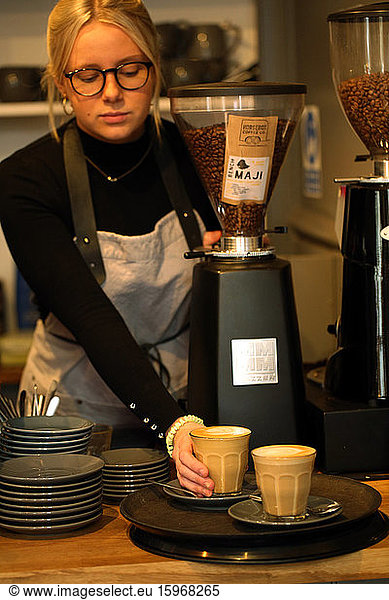 Blond woman wearing glasses and apron standing at counter in a cafe  placing two cafe lattes on a tray.