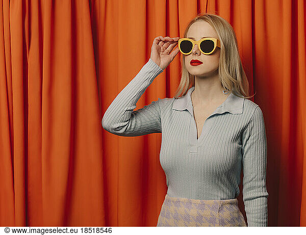 Blond woman wearing sunglasses standing in front of orange curtain