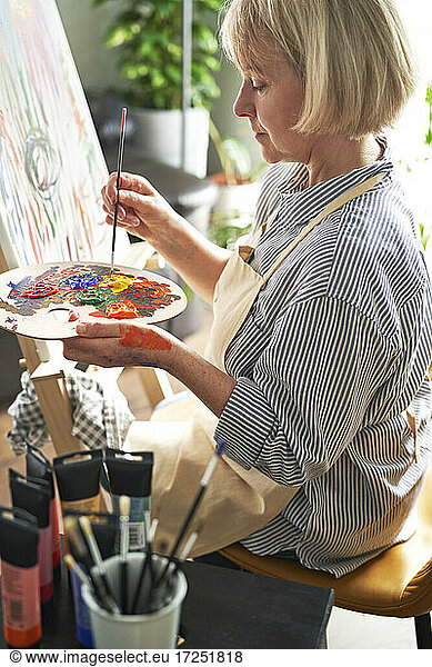 Blond woman with paintbrush mixing colors on palette in front of easel at home
