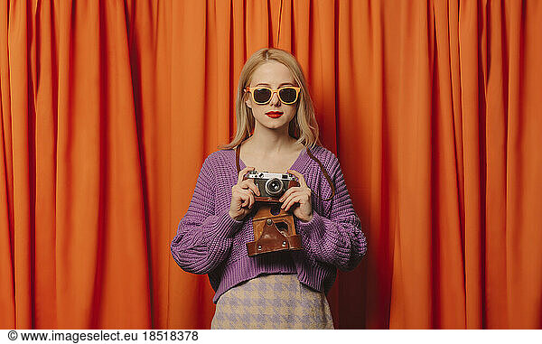 Blond woman with vintage camera standing in front of orange curtain
