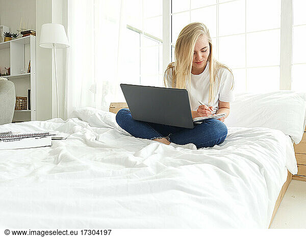 blond young female making notes with pen with latop on her nees in bed