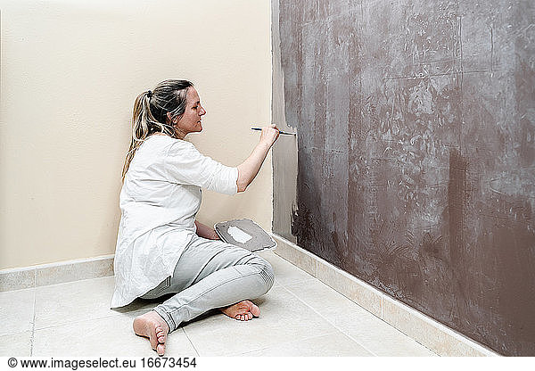 blonde barefoot woman sitting on her floor starts painting a brown wall into a grey color with a small paintbrush. The woman wears a white shirt and a grey trousers. Horizontal photo.