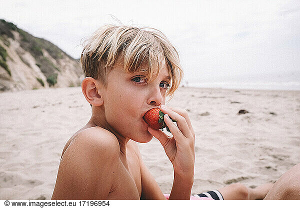 Blonde Boy Eats a Strawberry on a Sandy Beach