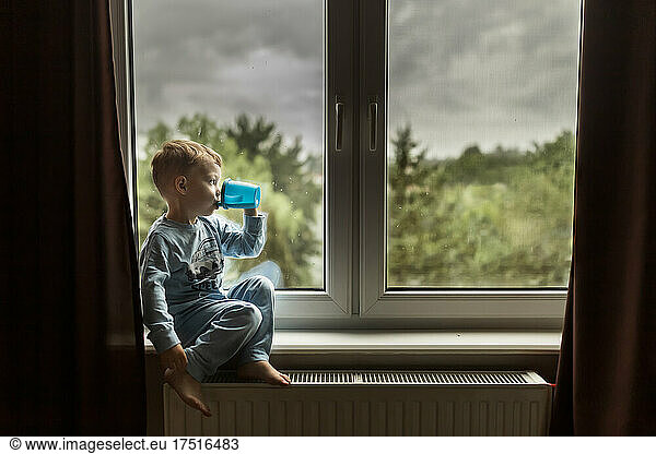 Blonde boy in blue pajamas drinking milk and sitting on windowsill