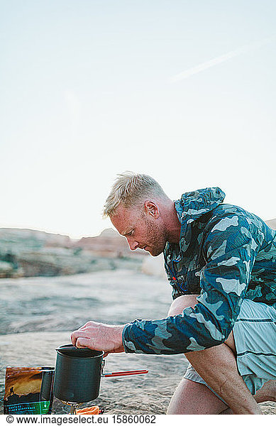 blonde camper with scruff and camo jacket boils water for dinner