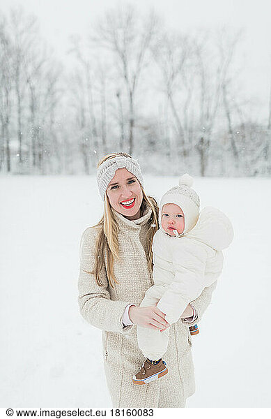 Blonde  Caucasian mother holds bundled-up baby in snow.