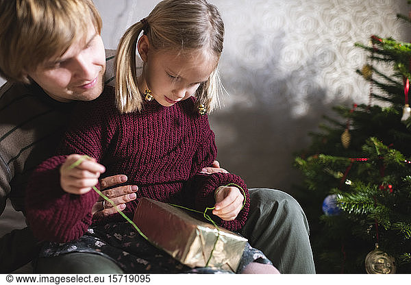 Blonde girl sitting on father's lap and opening Christmas present