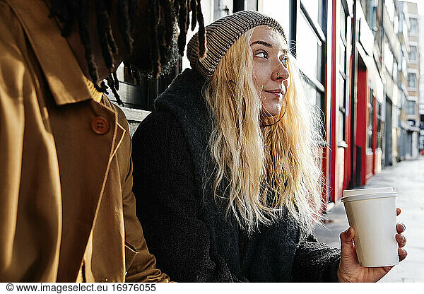 blonde young female sitting outside while drinking coffee with her friend. multicultural friendship