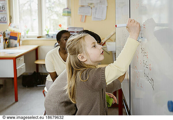 Blondes Mädchen löst ein Matheproblem  während sie auf die Tafel im Klassenzimmer schreibt