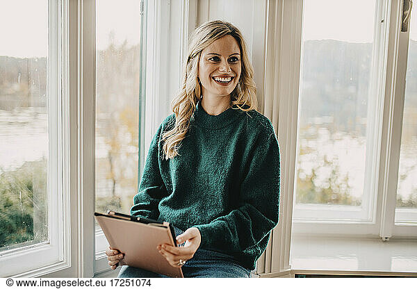 Blondhaarige Frau in Sweatshirt sitzt mit Tablet am Fenster im Büro