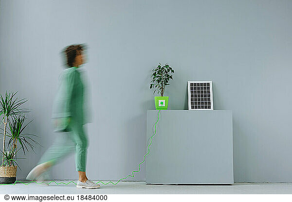 Blurred woman walking by electric plug connected to potted plant near solar panel in front of gray wall