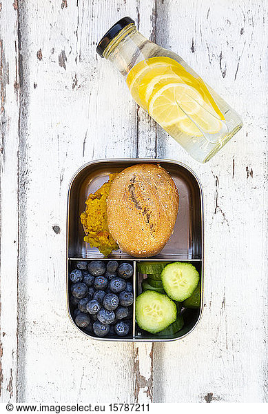 Bottle of lemonade and lunch box with cucumber slices  blueberries and bun with lentil paste