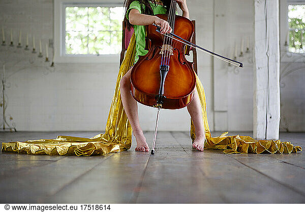 Bottom-half of barefoot child in gold cape playing cello in empty room