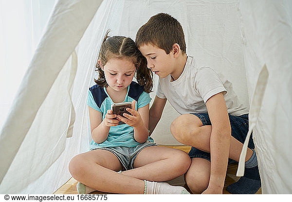 Boy and girl looking at a smartphone inside a white teepee tent inside their house. Technology concept