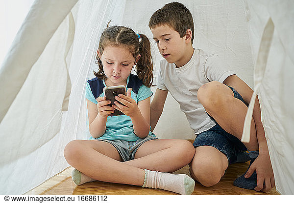 Boy and girl looking at a smartphone inside a white teepee tent inside their house. Technology concept