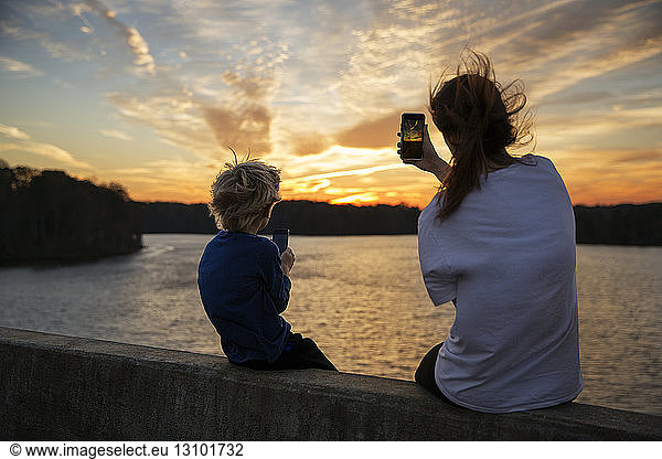 Boy and girl sitting on ledge photographing sunset