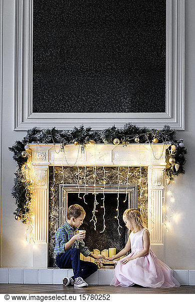 Boy and his little sister sitting together in front of fireplace at Christmas time