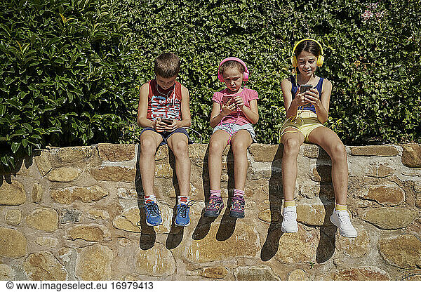 Boy and two girls looking at their smart phones and listening to music with headphones sitting on a stone wall in sunny day. Technology concept