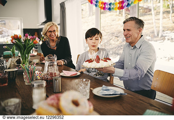 Boy blowing candles on birthday cake while sitting with grandparents at table