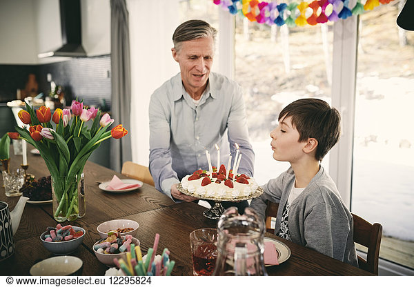 Boy blowing candles while grandfather holding birthday cake at table during party