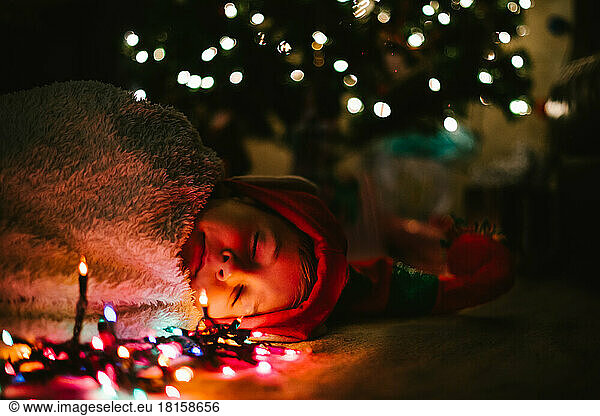 Boy child sleeps in cozy blanket and elf hat under Christmas tree