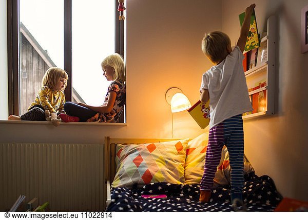 Boy choosing books from shelf with sisters sitting on window sill at home