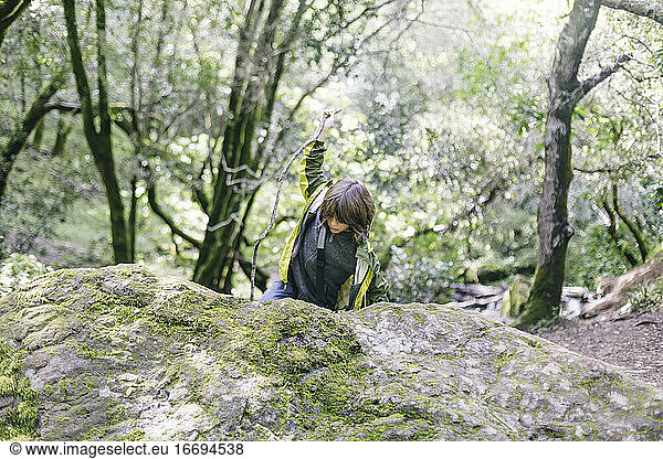 Boy climbing up rock with walking stick by Forest