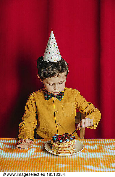 Boy cutting birthday at table in front of red curtain