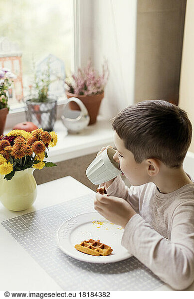 Boy drinking hot chocolate from cup on dining table in kitchen