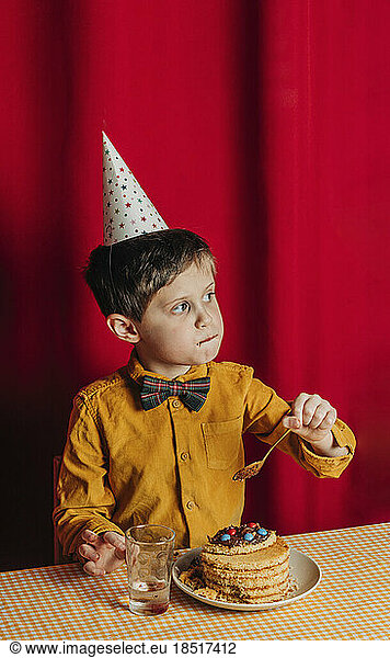 Boy eating birthday cake at table in front of red curtain