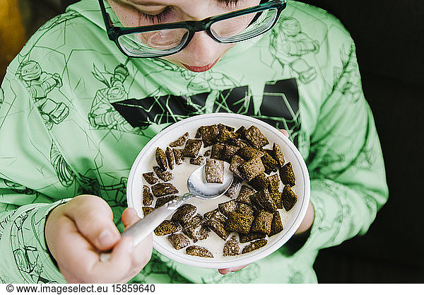 Boy eating chocolate cereal from a white bowl.