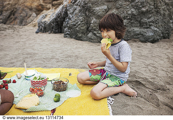 Boy eating taco while sitting on sand at beach