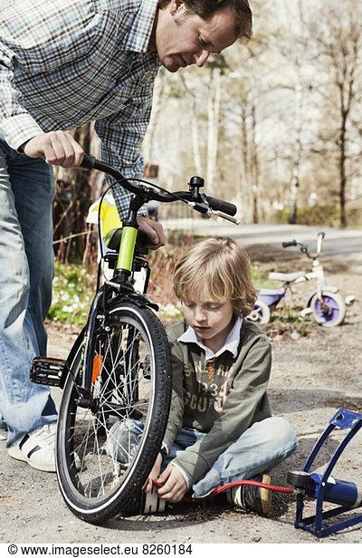 Boy filling bicycle tire with foot pump while father watching him on road