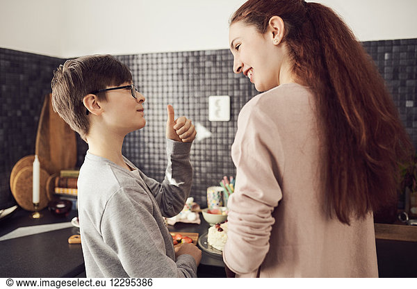 Boy gesturing thumbs up to sister while preparing food at kitchen