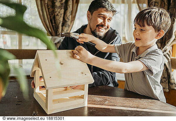Boy hammering nail on birdhouse by father at home
