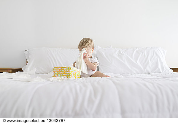 Boy hiding face with facial tissue while playing on bed at home