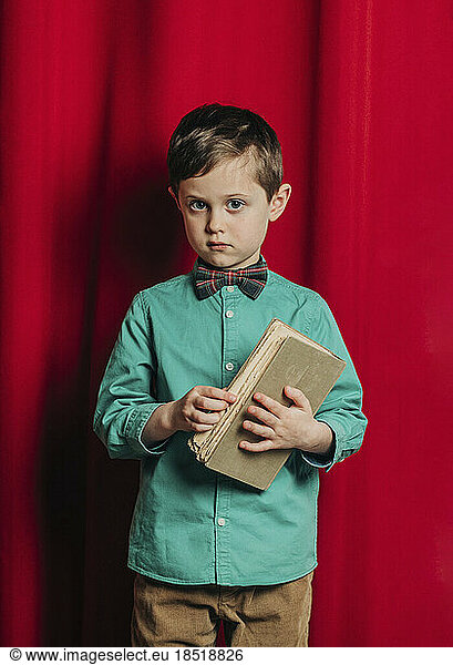 Boy holding book standing in front of red curtain