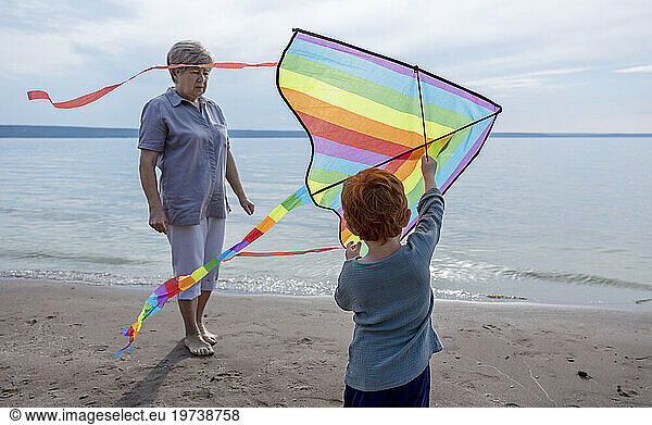 Boy holding colorful kite with grandmother near shore at beach