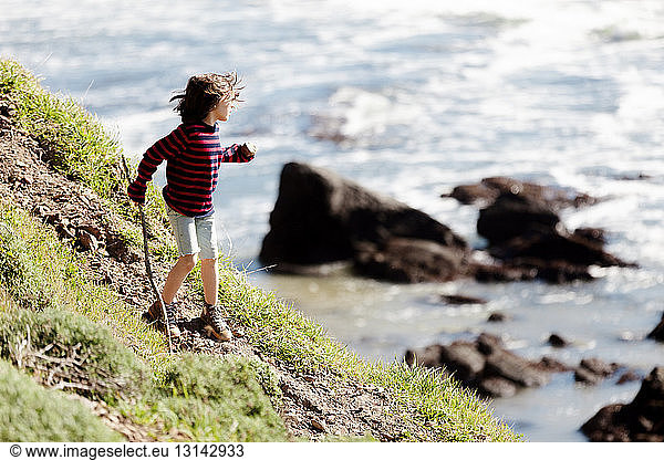 Boy holding stick climbing mountain by sea during sunny day