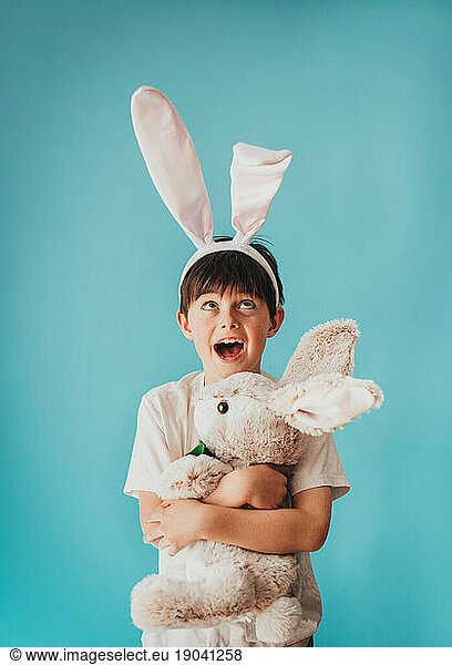Boy holding stuffed rabbit looking up at bunny ears on his head.