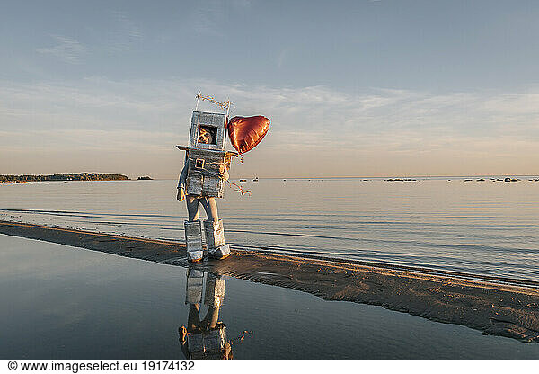 Boy in astronaut suit with heart shape balloon on beach at sunset